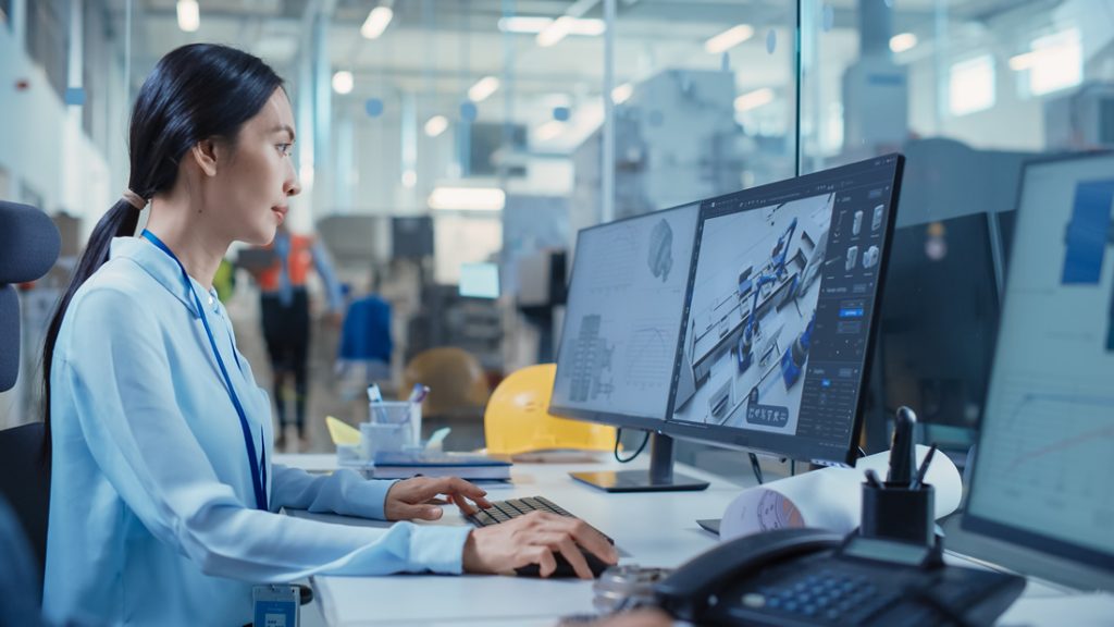 Person an einem CAD-Arbeitsplatz in industrieller Umgebung, die auf zwei Monitoren ein 3D-Modell und technische Konstruktionsdaten bearbeitet.